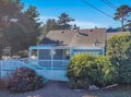 Charming coastal cottage with covered deck surrounded by mature trees and flowering gardens in peaceful neighborhood setting.