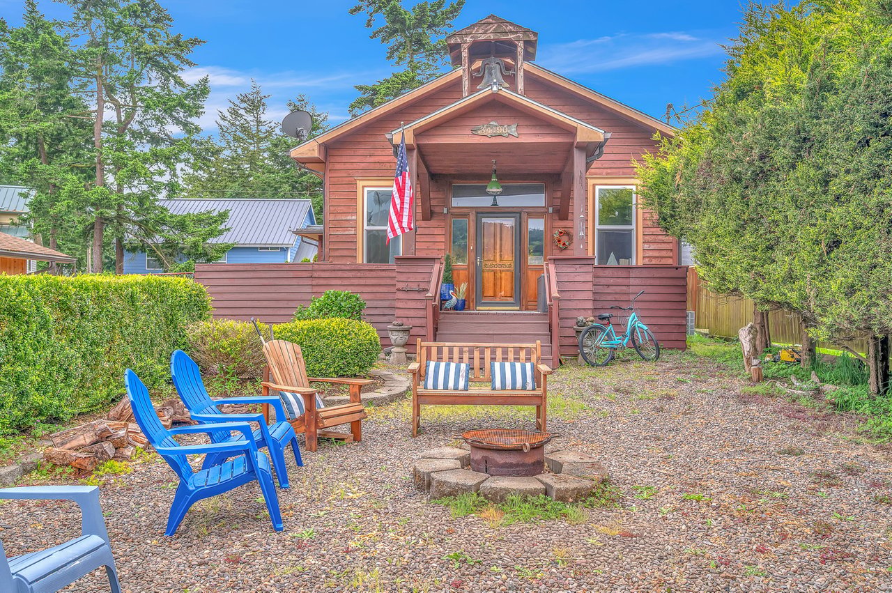 Charming red cottage with a welcoming front porch and fire pit seating area, surrounded by mature trees in a peaceful neighborhood setting.