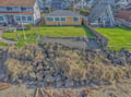 Aerial view showcases the beachfront property's coastal location among charming neighborhood homes with natural rock shoreline and green lawns.