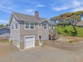 Coastal cottage with two-story design, garage parking, wooden deck, and lush natural landscaping surrounded by mature trees.