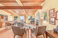 Unwind in your open-concept living area where warm wood beams frame stunning water views through every window. Gather by the fireplace or relax in wicker chairs while the coastline stretches just beyond your door.