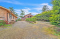 A peaceful property surrounded by mature evergreens and manicured hedges, with ample parking and multiple buildings set on a spacious gravel lot beneath bright blue skies.