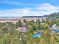 Aerial view of a coastal vacation rental nestled among evergreens, with expansive sandy beach and ocean bay stretching into the distance beneath open skies.