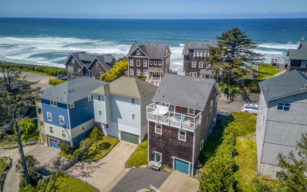 Aerial view of a charming coastal neighborhood featuring multi-story beach homes nestled among mature trees, with the Pacific Ocean waves rolling in just steps away from the property.