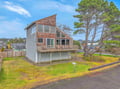 Modern coastal home with wood shingle accents and expansive windows, nestled among mature pine trees in a quiet residential neighborhood.