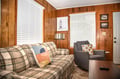 Sink into the plaid sofa or curl up in the cozy gray chair—your living room welcomes you with warm wood paneling, natural light, and a relaxed, cabin-like charm.