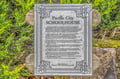 A historic marker commemorating the Pacific City Schoolhouse, mounted on weathered wood with decorative metalwork and framed by vibrant greenery.