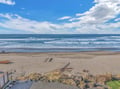 Pristine sandy beach stretches along the coastline with gentle waves rolling onto shore under a bright blue sky dotted with white clouds.