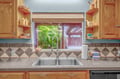 Prepare your favorite meals at the double-basin sink, surrounded by warm wood cabinetry and charming tile backsplash, as natural light streams through the window overlooking lush greenery.