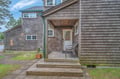 Charming cedar-shingled cottage featuring a welcoming covered porch with comfortable seating, surrounded by natural coastal landscaping and mature trees.