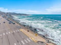 Scenic coastal road runs alongside dramatic ocean waves and rocky shoreline with mountain backdrop.