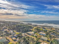 Coastal community nestled between rolling hills and ocean waves, with residential neighborhoods leading to sandy shores and dramatic cloudy skies.