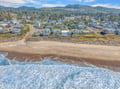 Aerial view of beachfront community with ocean waves meeting sandy shore, surrounded by forested coastal mountains.