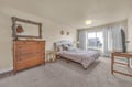 Relax in your spacious bedroom featuring warm wood furnishings, soft carpeting, and natural light streaming through white curtains.