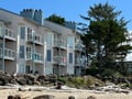 Beachfront property with private balconies, nestled between mature evergreens and sandy shoreline. The building sits along a quiet stretch of beach near the water's edge.
