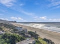 An elevated coastal view showcasing a wide sandy beach stretching along the Pacific shoreline, with rolling waves and nearby coastal homes nestled among lush hillside greenery under bright blue skies.