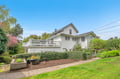 Charming white weatherboard home with wraparound deck sits among manicured gardens in peaceful residential setting.