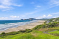 A stunning coastal landscape featuring a wide sandy beach, rolling green hills, and ocean views stretching toward distant bluffs and the horizon.