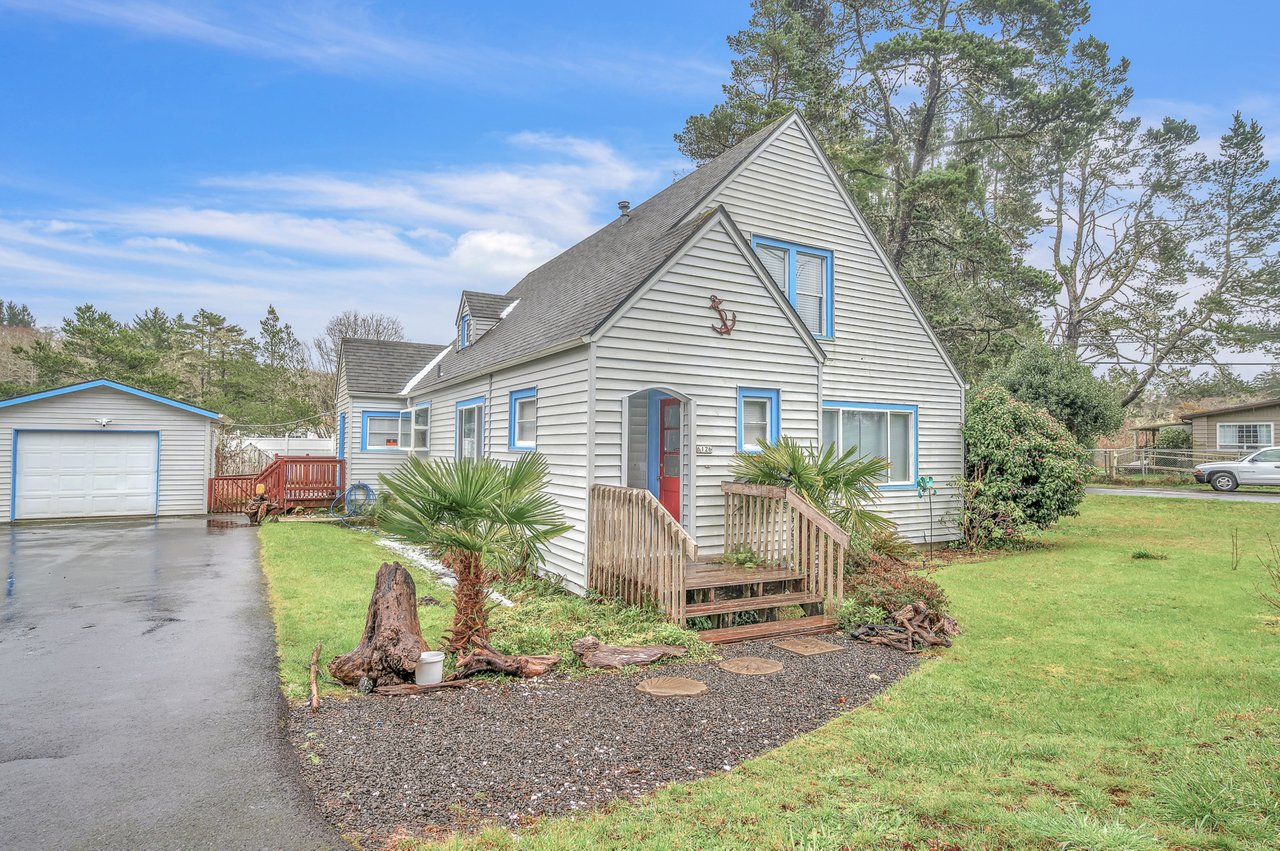 Charming coastal cottage with blue trim sits among towering trees, featuring a welcoming front deck and mature landscaping in a peaceful residential setting.