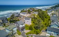 Aerial view of a coastal neighborhood with residential properties situated along a scenic stretch of sandy beach and ocean waves, surrounded by lush greenery and coastal landscape.