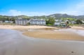 This aerial view captures the beachfront community nestled along the sandy shore, with forested hills rising in the background and calm waters stretching before the property.