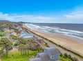Aerial view of a coastal neighborhood nestled between rolling hills and a pristine sandy beach, where gentle waves meet the shore beneath an expansive blue sky.