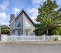 A charming gray A-frame cottage with crisp white trim and picket fence stands ready to welcome guests to their coastal getaway.