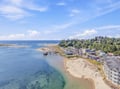 An aerial view captures the coastal town nestled between ocean waters and forested hillside, showcasing the beachfront area and nearby community during a bright summer day.