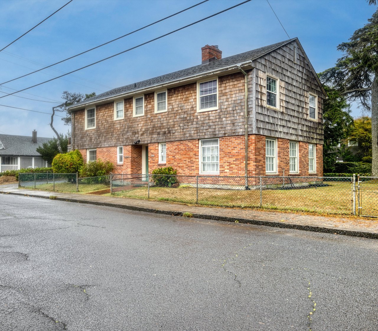 A charming two-story home featuring classic shingled upper level and brick foundation, nestled in a peaceful residential neighborhood with mature trees and a spacious fenced yard.