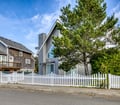 A charming gray beach cottage with distinctive A-frame architecture, surrounded by mature pines and enclosed by a crisp white picket fence in a coastal neighborhood.