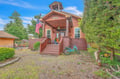 Charming red schoolhouse-style cottage features a bell tower, covered porch, and vibrant blue bike, set amidst lush greenery for a welcoming arrival.