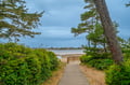A peaceful pathway leads through coastal greenery to a sandy beach and calm bay, framed by towering evergreens and lush vegetation—part of the natural landscape surrounding the property.
