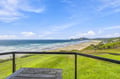 This stunning coastal property overlooks a pristine sandy beach framed by rolling waves and lush green headlands stretching into the distance beneath an endless blue sky.