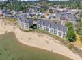Aerial view of a beachfront property nestled in a charming coastal town, with forested hills and a quiet neighborhood setting.