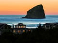 Dramatic coastal sunset paints the sky in warm hues above a striking sea stack and beachfront homes.