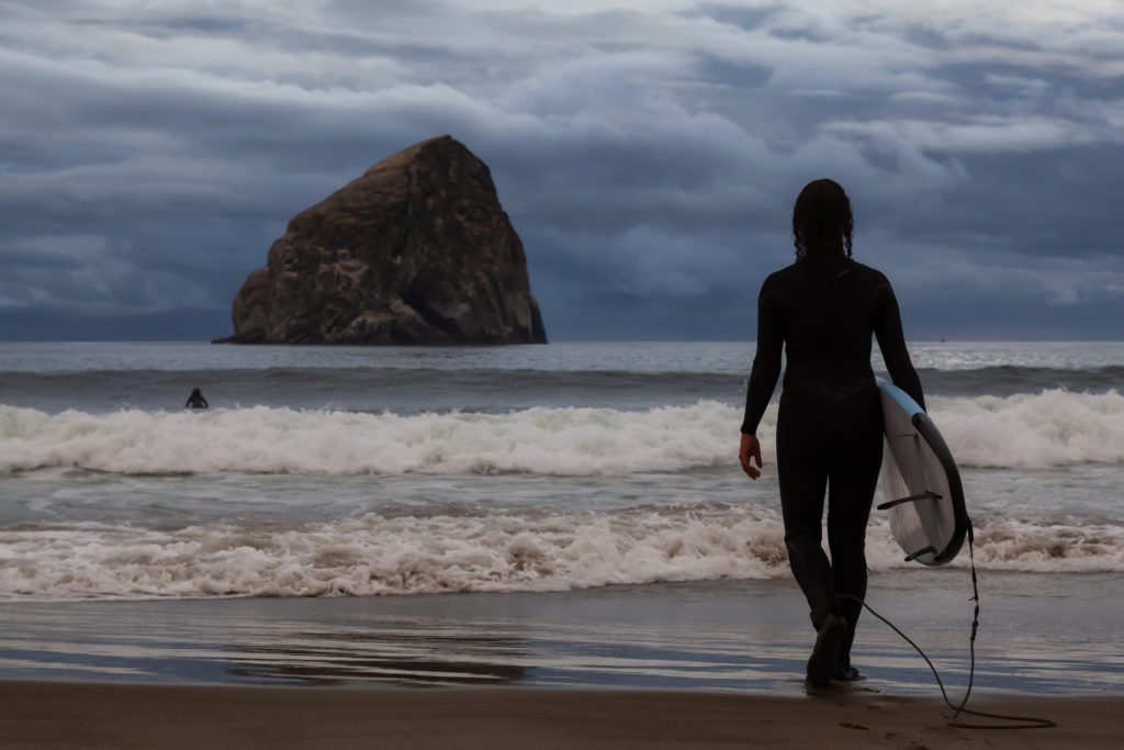 Adventurous Girl with a Surf Board is going surfing in the Ocean during a cloudy summer sunset. Taken in Pacific City, Oregon Coast, United States of America.